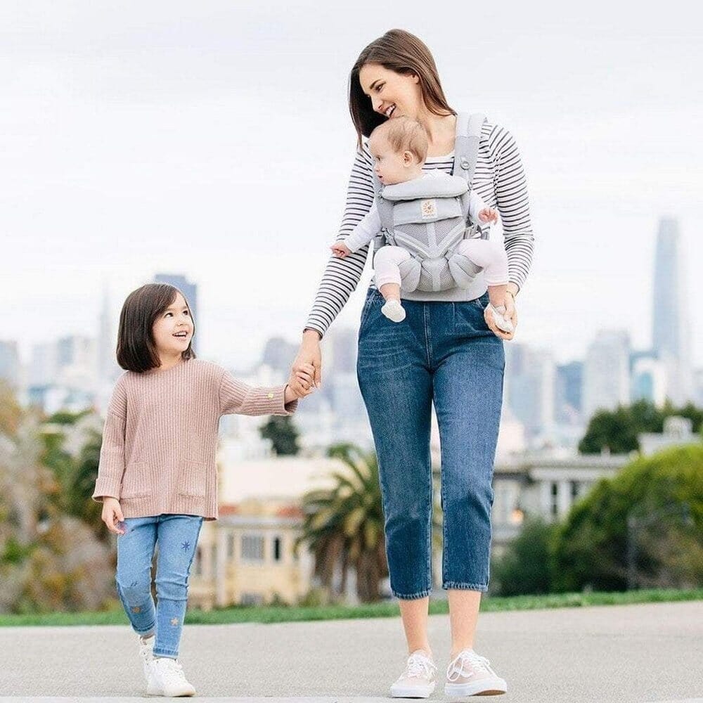 Porte Bébé Deux Coeur - maman marche avec bébé et tient la main de sa fille. Confort et liberté pour toute la famille.